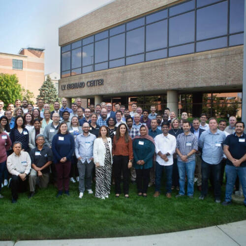 faculty and staff stand together on a green lawn in front of WM EBERHARD CENTER. They are arranged in several rows, with some people standing and a few seated in front.